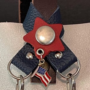 May include: Close-up of a leather accessory featuring a red star with a silver button, blue leather accents, and a small American flag charm. Includes silver key rings and clasps. The design incorporates patriotic colors and elements.