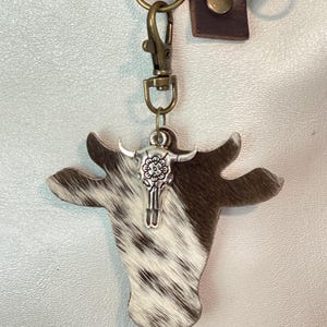 May include: A cowhide keychain featuring a cow head shape with a silver-colored steer skull charm. The keychain has a brown leather strap and a metal clasp for attaching to keys or a bag. The cow head has a brown and white spotted pattern.