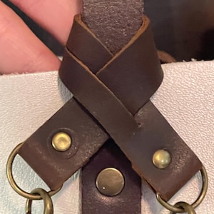 May include: Close-up of a brown leather strap detail with brass-coloured hardware. The strap is knotted and features two snap closures and two metal clasps. The leather has a textured appearance.