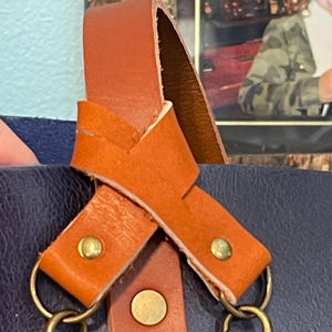 May include: A close-up of a brown leather strap with two brass clasps. The strap is crossed over itself and has two brass rivets on the strap.