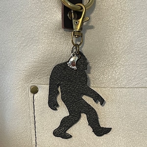 May include: A black leather Bigfoot keyring with a silver charm attached to a brass key ring.