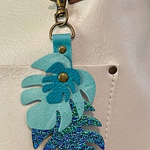 May include: A teal leather keychain featuring a monstera leaf design. The leaf has a textured surface with a smaller leaf detail and a brass-colored rivet. The keychain includes a clasp and a leather strap.
