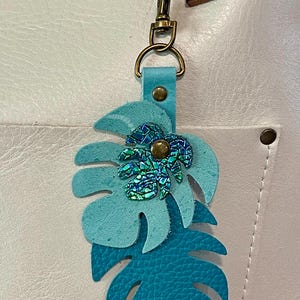May include: A turquoise leather keychain featuring a layered monstera leaf design. The top leaf has a textured, iridescent embellishment. The keychain includes a brass-colored clasp and a blue leather strap.