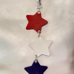 May include: A patriotic keychain featuring three leather stars in red, white, and blue. The stars are connected by small metal rings and attached to a silver-colored clasp. A small American flag charm is also included.