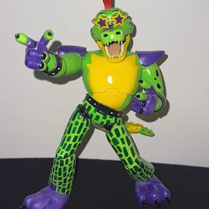 Op de afbeelding: Een antropomorfe alligator-actiefiguur met een groene huid, een gele borstplaat en paarse accenten. Het figuur draagt een stervormige zonnebril, heeft een rode hanenkam en een groen-zwart ruitpatroon op de benen en de staart.