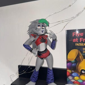 May include: A gray figurine of a wolf-like character with a red top, purple accents, and a green visor. The figure is posed on a black shelf, with a book visible in the background. The figurine is from the Five Nights at Freddy's franchise.