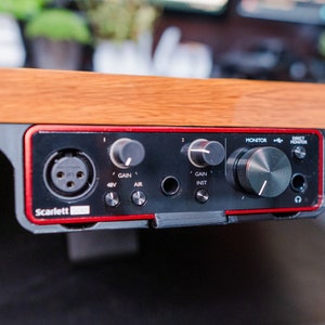 Peut inclure: Une interface audio Focusrite Scarlett 2i2 noire avec des accents rouges. Elle possède deux boutons de gain, un pour le moniteur et un pour le moniteur direct. Elle possède un bouton 48V, un bouton air et un bouton inst. Elle possède une prise jack 1/4 pouce et un connecteur XLR.