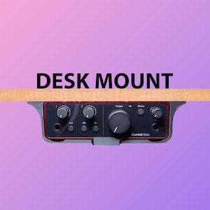 Può includere: Un'interfaccia audio nera e grigia con manopole e pulsanti, montata su una superficie da scrivania in legno. Il testo "Scarlett Solo" è stampato sulla parte anteriore del dispositivo. Il testo "DESK MOUNT" è stampato sopra il dispositivo.