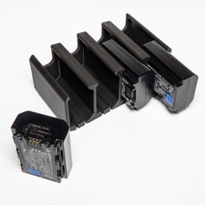 Könnte beinhalten: Ein schwarzer Batteriehalter aus Kunststoff mit vier Steckplätzen zum Laden von Kamerabatterien. Drei Batterien befinden sich im Halter, eine ist separat.