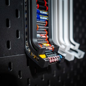 Könnte beinhalten: Ein schwarzer Batterie-Organizer aus Kunststoff mit einem weißen Etikett mit der Aufschrift "VARIA" und einer Reihe von AA-Batterien. Die Batterien sind vertikal angeordnet und werden durch einen federbelasteten Mechanismus gehalten.