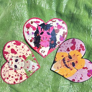Puede incluir: Tres pegatinas en forma de corazón con un fondo de pintura salpicada rosa, blanco y rojo. Cada pegatina presenta un personaje diferente de una película de terror. La primera pegatina presenta un personaje enmascarado de cerdo con cabello negro. La segunda pegatina presenta un personaje con una máscara de hockey. La tercera pegatina presenta un personaje con una máscara amarilla y negra y cabello rubio.