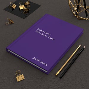 May include: A purple journal with the text "Notes from The Crazy Train" and "Jackie Smith" printed in white. Gold and black pencils and gold binder clips are arranged around the journal on a dark gray surface.