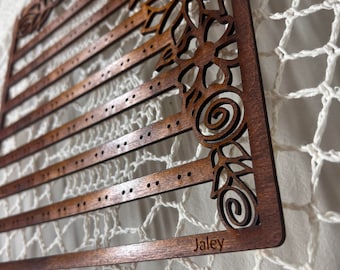 May include: Wooden earring holder with five rows of holes and a floral design on the right side. The holder is stained a dark brown color and has the name "Jaley" engraved on the bottom.