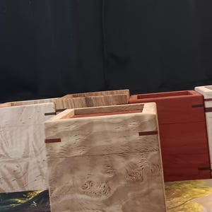May include: Wooden boxes with a light wood grain pattern, accented with red trim. The boxes are rectangular and appear to be handmade. A few playing cards and a die are visible in the foreground, suggesting a game-related theme.