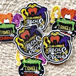 Puede incluir: Una colección de parches bordados con dos diseños distintos. Un diseño muestra un caldero negro con líquido verde y el texto I SMELL CHILDREN. El otro diseño presenta tres siluetas de cabello estilizadas y el texto IT'S ALL A BUNCH OF HOCUS POCUS.