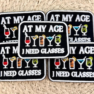 May include: Five rectangular black embroidered patches featuring the white text AT MY AGE I NEED GLASSES. Each patch displays a row of five colourful cocktail glasses in red, yellow, orange, blue, and green, with white stitching around the edges.