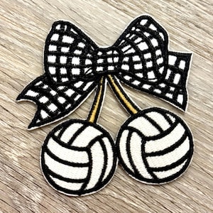 Könnte beinhalten: Gestickter Aufnäher mit einer schwarz-weiß karierten Schleife über zwei weißen Volleyballen mit schwarzen Details. Die Volleybälle sind mit goldenen Stielen an der Schleife befestigt. Der Aufnäher befindet sich auf einer Holzoberfläche.
