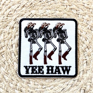 May include: Square patch featuring three skeleton cowboys wearing hats and boots. The skeletons are black and white, with brown boots and hats. The text "YEE HAW" is in black, below the skeletons.