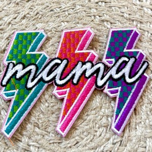 May include: Three embroidered lightning bolt patches in teal, orange, and purple, each with a checkered pattern. The word "mama" is embroidered in white script across the center of the patches. The patches are on a woven background.