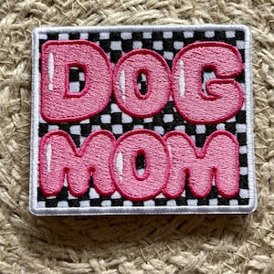 May include: Embroidered patch with the words "DOG MOM" in pink bubble letters. The text is outlined in white and set against a black and white checkered background. The patch has a white border.