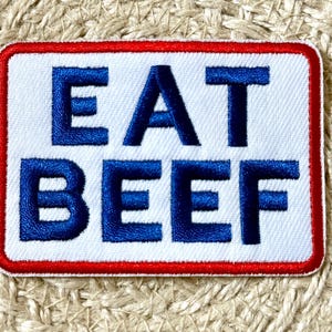 May include: Embroidered patch with the words "EAT BEEF" in bold, blue letters on a white background. The patch has a red border and is rectangular in shape. The patch is on a textured, beige surface.