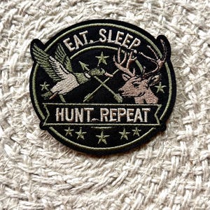 Könnte beinhalten: Ein runder gestickter Aufnäher mit schwarzem Hintergrund und olivgrünem Rand. Er zeigt eine fliegende Ente, einen Hirschkopf und zwei gekreuzte Pfeile. Der Text lautet EAT. SLEEP oben und HUNT. REPEAT auf einem Banner, mit kleinen Sternen.