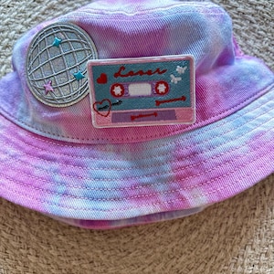 Puede incluir: Un gorro de cubo de color rosa y azul tie-dye con una bola de discoteca plateada y un parche de cinta de casete rosa y azul. El parche de cinta de casete tiene la palabra "lover" escrita en él.