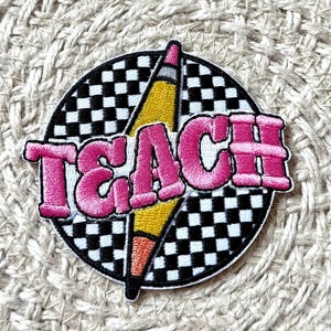 May include: Embroidered patch featuring the word "TEACH" in pink, outlined in white, over a yellow pencil. The patch is set against a black and white chequered background, with a black border.