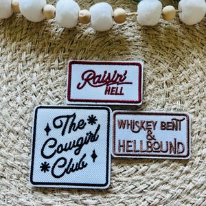 May include: Three embroidered patches with text. The top patch is rectangular with the words "Raisin' Hell" in red script. The middle patch is square with "The Cowgirl Club" in black script. The bottom patch reads "Whiskey Bent & Hellbound".