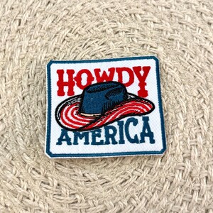 May include: A square embroidered patch with a white background and a dark blue border. The text HOWDY is stitched in red above a dark blue cowboy hat with a red and white striped brim. The word AMERICA is stitched in dark blue below the hat.
