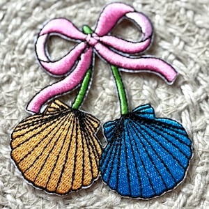 May include: Embroidered patch featuring two seashell designs, one gold and one blue, connected by green stems and a pink bow. The seashells have detailed black outlines, and the bow has a pink outline. The patch is on a textured, neutral background.