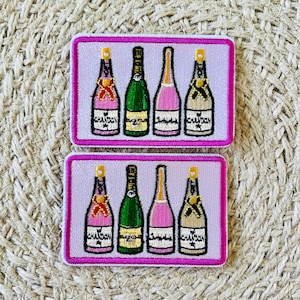 Puede incluir: Dos parches bordados en rosa y blanco con seis botellas de champán con etiquetas de lámina dorada. Las botellas están dispuestas en dos filas de tres. Los parches son perfectos para añadir un toque de brillo a cualquier proyecto.