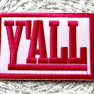 May include: Embroidered patch with the word "Y'ALL" in bold, red letters. The letters are stitched onto a white background, framed by a red border. The patch is rectangular and appears to be made of fabric.