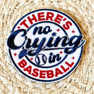 May include: A white embroidered patch with blue and red stitching. The patch is round and features a baseball with the text "There's no crying in baseball".