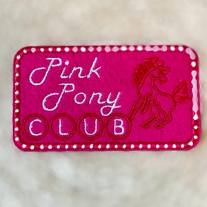 Op de afbeelding: Een rechthoekige, felroze patch met witte geborduurde tekst die luidt "Pink Pony CLUB". Een rood pony-silhouet staat rechts. De patch is omrand met witte stippen en een witte rand.