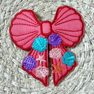 May include: A pink fabric bow with a seashell theme. The bow features a detailed design with a red outline, and is decorated with turquoise, purple, and pink seashell-shaped embellishments, along with starfish.