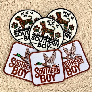 Pode incluir: Uma coleção de emblemas bordados com três designs circulares apresentando um cão camuflado e pegadas, e três designs quadrados com um pato-real em voo. Todos os emblemas incluem o texto SOUTHERN BOY em letras pretas ou castanhas.
