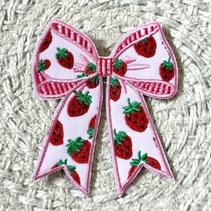 Könnte beinhalten: Ein pinkfarbener Aufnäher in Schleifenform mit Erdbeermuster. Der Bogen hat einen weißen Hintergrund mit roten Erdbeeren und grünen Blättern. Die Ränder sind pink umrandet.