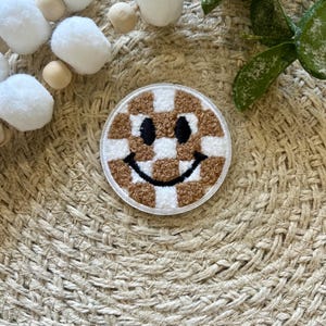 Op de afbeelding: Een bruin en wit geruite smiley-face patch met een zwarte omtrek. De patch is gemaakt van pluche stof en ligt op een geweven oppervlak.