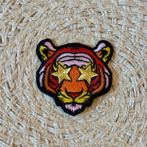 May include: Embroidered tiger patch with star-shaped eyes. The patch features a black border, with orange, red, pink, and yellow thread. The patch is on a woven, beige surface.
