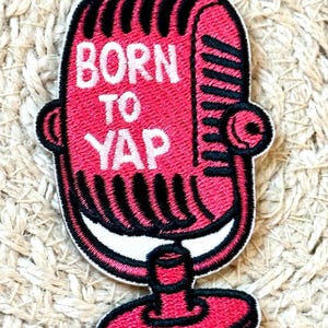 Könnte beinhalten: Ein pinkfarbener, gestickter Aufnäher in Form eines Vintage-Mikrofons. Der Aufnäher zeigt die Worte "BORN TO YAP" in weißer Schrift. Das Mikrofon hat schwarze Details und eine weiße Umrandung, vor einem strukturierten Hintergrund.