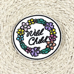 May include: White embroidered patch with a floral wreath and the text "Wild Child" in black script.