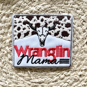 May include: A white embroidered patch with a brown and white cow skull design. The text "Wranglin' Mama" is in red and black lettering.