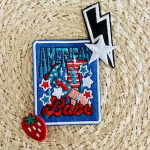 Op de afbeelding: Een verzameling geborduurde patches met patriottische ontwerpen. Bevat een blauwe patch met de tekst "American Babe" en een afbeelding van een vrouwenbeen, een aardbei, een witte ster en een zwart-witte bliksem.