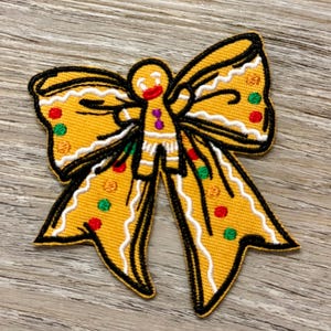 以下が含まれることがあります： ジンジャーブレッドマンのリボンの形の刺繍パッチ。 黄色のリボンは黒いアウトラインと白いトリムがあり、赤、緑、オレンジの円形のアクセントがあります。 ジンジャーブレッドマンは笑顔の顔と紫色のアクセントがあります。
