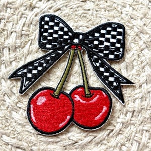 May include: Embroidered patch featuring two red cherries with green stems and a black and white checkered bow. The patch has a white border and is set against a textured, beige background. This patch is suitable for clothing and accessories.