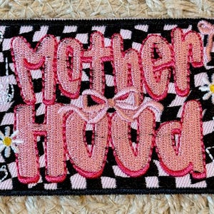 May include: Embroidered patch with the word "Motherhood" in pink, outlined in red, on a black and white checkered background. The patch features bows, daisies, and other decorative elements.