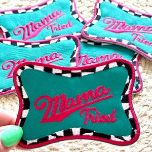 以下が含まれることがあります： 「ママトライ」とピンクの刺繍が施された、ターコイズブルーと白のチェック柄の布製パッチが4つあります。