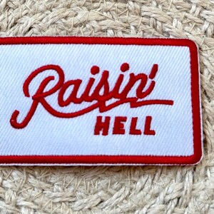 Op de afbeelding: Geborduurde patch met een witte achtergrond en een rode rand. De patch bevat de woorden "Raisin' HELL" in rode, gestileerde letters. De patch is rechthoekig.