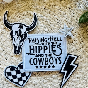 Puede incluir: Un conjunto de parches para planchar que incluyen una calavera, un corazón con un patrón de cuadros, un rayo, una estrella y un parche blanco con el texto "Raising Hell with the Hippies and the Cowboys" en negro.
